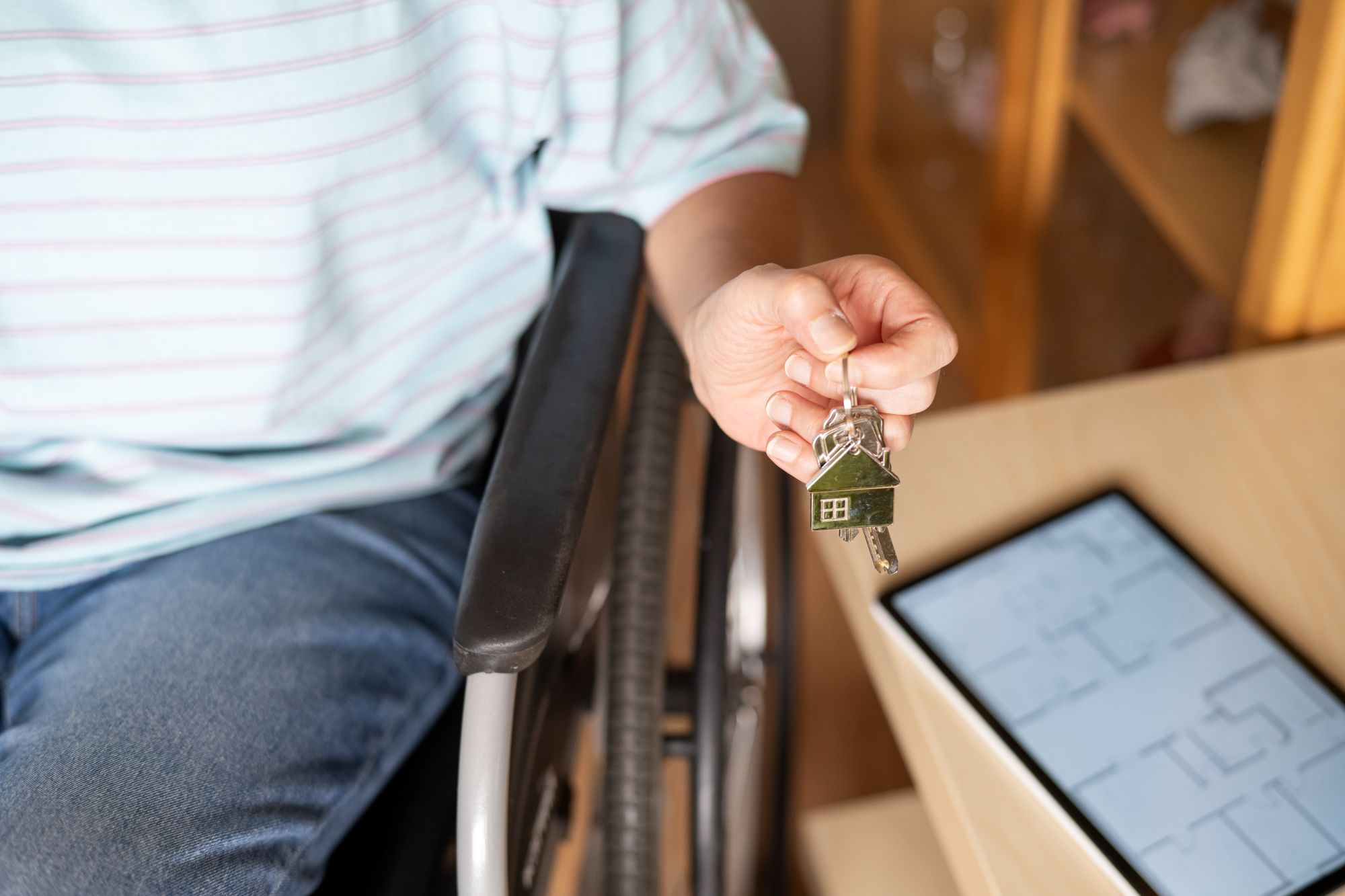 Person in a wheelchair holding keys to a new home with blueprint in the background